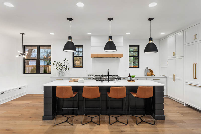 Modern kitchen with a large white island, four brown barstools, black pendant lights, white cabinets, wood floor, and black-framed windows letting in natural light.